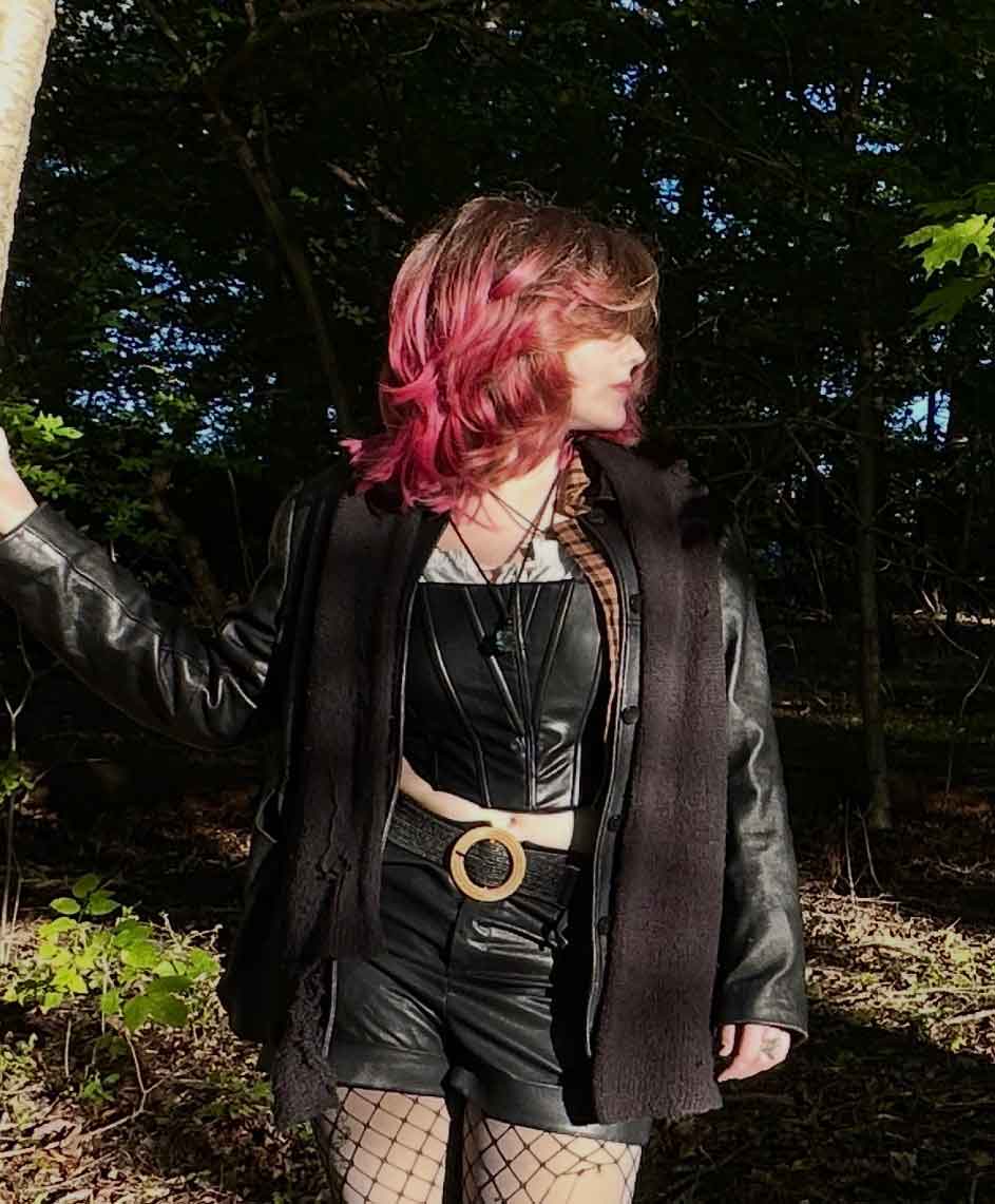 A person with pink-tinted hair and tattoos from a studio in North Wales stands in a forest wearing a black corset, shorts, fishnet stockings, and a black jacket.